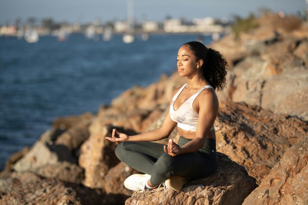 Woman meditating on rocks by the ocean, embodying calmness and wellness.